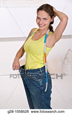 Stock Photograph - young woman in too big jeans after slimming with a measuring tapa around her neck, smiling looking at camera. Fotosearch