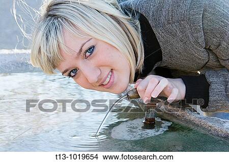 Picture - Young woman is drinking running fresh water from small fountain pipe. Fotosearch