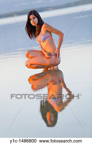 Stock Image - Young woman is squatting in a shallow water tying her swimsuit panties. Fotosearch