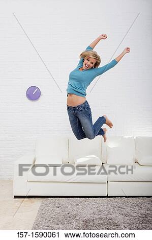 Stock Image - Young woman jumping for joy off her sofa. Fotosearch
