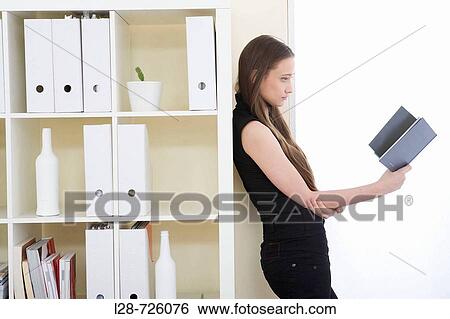 Young woman leaning against shelves and reading book View Large Photo Image Stock Photograph - Young woman leaning against shelves and reading book. Fotosearch