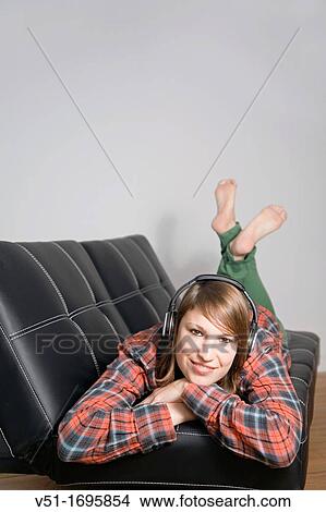 Picture - Young woman listening to music on the couch smiling at camera. Fotosearch