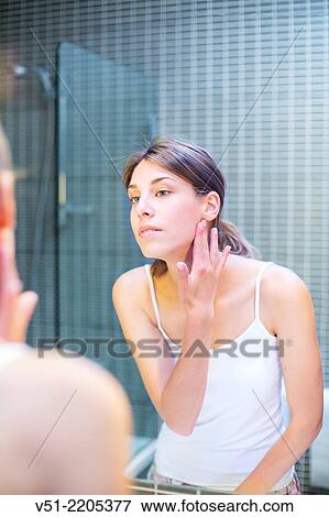Stock Photo - Young woman looking at herself in the mirror in the bathroom. Fotosearch