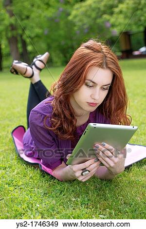 Young woman lying on the grass in a park and reading on a tablet pc View Large Photo Image Stock Photo - Young woman lying on the grass in a park and reading on a tablet pc. Fotosearch