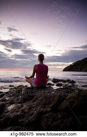Stock Photography - Young woman meditating on rocks at sunset in front of ocean surf at Playas del Coco, Costa Rica. Fotosearch