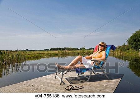 Stock Photograph - Young woman relaxing in a deck chair at a pond. Fotosearch