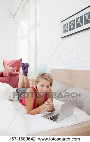 Young woman relaxing on her bed on a laptop View Large Photo Image Stock Image - Young woman relaxing on her bed on a laptop. Fotosearch