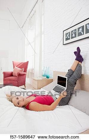 Young woman relaxing on her bed on her laptop View Large Photo Image Stock Photo - Young woman relaxing on her bed on her laptop. Fotosearch