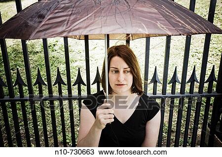 Young woman, sitting in front of an iron fence, holding an umbrella. View Large Photo Image Stock Image - Young woman, sitting in front of an iron fence, holding an umbrella.. Fotosearch