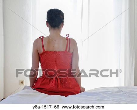 Stock Photograph - Young woman sitting on bed, rear view. Fotosearch