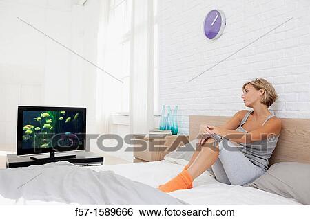 Stock Photograph - Young woman sitting on her bed watching TV. Fotosearch