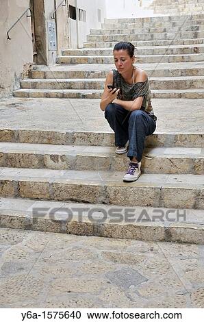 Stock Image - Young woman sitting on stairs outdoors text messaging. Fotosearch