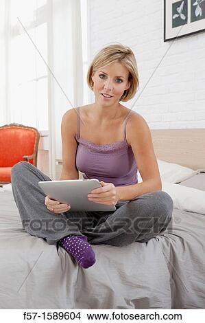 Young woman sitting on the edge of her bed with a tablet computer View Large Photo Image Picture - Young woman sitting on the edge of her bed with a tablet computer. Fotosearch