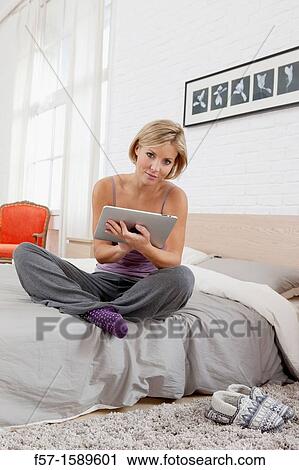 Young woman sitting on the edge of her bed with a tablet computer View Large Photo Image Stock Image - Young woman sitting on the edge of her bed with a tablet computer. Fotosearch