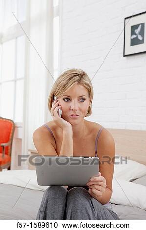 Young woman sitting on the edge of her bed with a tablet computer and talking on her mobile phone View Large Photo Image Stock Image - Young woman sitting on the edge of her bed with a tablet computer and talking on her mobile phone. Fotosearch