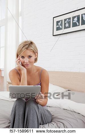 Young woman sitting on the edge of her bed with a tablet computer and talking on her mobile phone View Large Photo Image Stock Image - Young woman sitting on the edge of her bed with a tablet computer and talking on her mobile phone. Fotosearch
