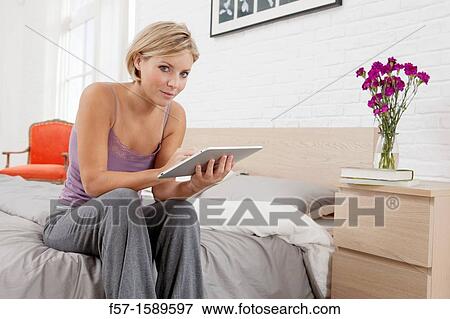 Young woman sitting on the edge of her bed with a tablet computer View Large Photo Image Stock Photo - Young woman sitting on the edge of her bed with a tablet computer. Fotosearch