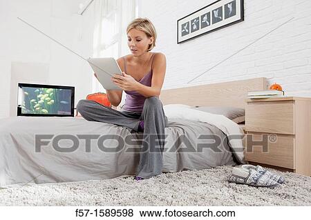 Young woman sitting on the edge of her bed with a tablet computer View Large Photo Image Stock Photo - Young woman sitting on the edge of her bed with a tablet computer. Fotosearch