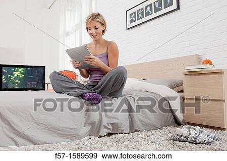 Young woman sitting on the edge of her bed with a tablet computer View Large Photo Image Stock Photo - Young woman sitting on the edge of her bed with a tablet computer. Fotosearch