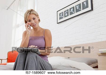 Young woman sitting on the edge of her bed with a tablet computer and talking on her mobile phone View Large Photo Image Stock Photo - Young woman sitting on the edge of her bed with a tablet computer and talking on her mobile phone. Fotosearch