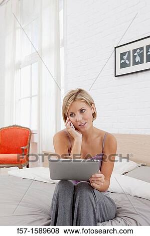 Young woman sitting on the edge of her bed with a tablet computer and talking on her mobile phone View Large Photo Image Stock Photo - Young woman sitting on the edge of her bed with a tablet computer and talking on her mobile phone. Fotosearch