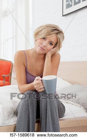 Young woman sitting on the edge of her bed drinking coffee View Large Photo Image Stock Photo - Young woman sitting on the edge of her bed drinking coffee. Fotosearch