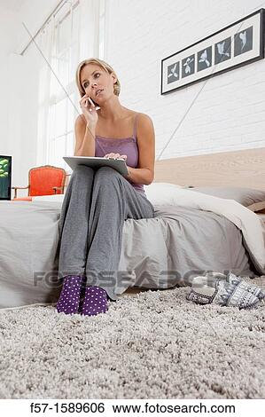 Young woman sitting on the edge of her bed with a tablet computer and talking on her mobile phone View Large Photo Image Stock Photograph - Young woman sitting on the edge of her bed with a tablet computer and talking on her mobile phone. Fotosearch