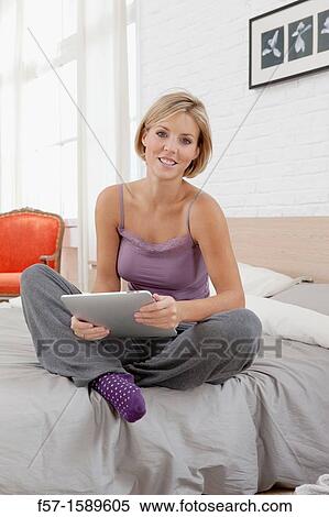 Young woman sitting on the edge of her bed with a tablet computer View Large Photo Image Stock Photography - Young woman sitting on the edge of her bed with a tablet computer. Fotosearch