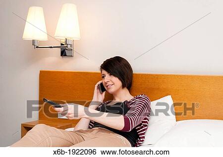 Stock Photograph - Young woman teenager sitting on bed watching TV and talking on mobile phone. Fotosearch
