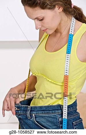 Stock Photo - young woman trying on an oversize jeans because she has slimmed after a diet with a measuring tape around her neck. Fotosearch
