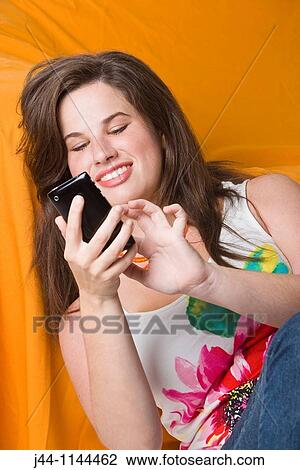 Stock Image - Young woman using mobile phone leaning on sofa smiling. Fotosearch