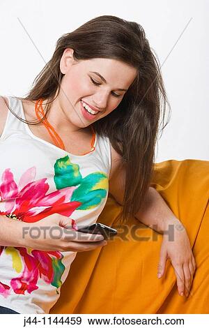 Young woman using mobile phone sitting on sofa smiling View Large Photo Image Stock Photo - Young woman using mobile phone sitting on sofa smiling. Fotosearch