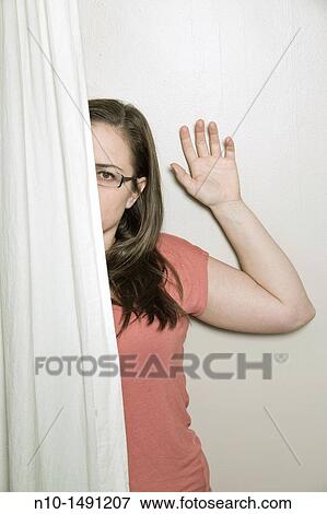Stock Photo - Young woman, wearing glasses, looking out from behind a curtain. Fotosearch