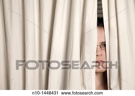 Stock Image - Young woman, wearing glasses, peeking out from behind two curtains. Fotosearch