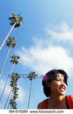 Young woman with flower in her hair smiling and looking out toward the unseen ocean with towering palm trees and blue sky behind her View Large Photo Image Stock Photo - Young woman with flower in her hair smiling and looking out toward the unseen ocean with towering palm trees and blue sky behind her. Fotosearch