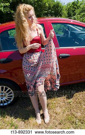Stock Image - Young woman with long curly hair is standing next to a Red car while holding a hem of her long skirt. Fotosearch