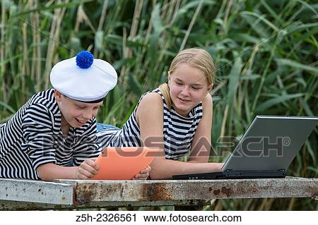 Stock Image - Boy and girl of twelve years in a sailor uniform with a laptop on the dock.. Fotosearch