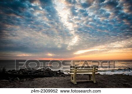 Stock Image - An empty bench sits on the edge of a beach at sunrise. Beautiful colors and dramatic clouds are in the distance. Jekyll Island, Georgia, USA. Fotosearch