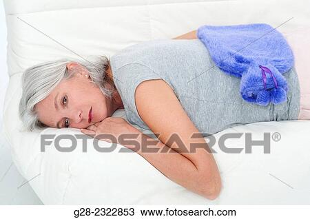 Stock Image - Beautiful mature woman, lying with a hot water bottle on her back, on a sofa bed. Fotosearch