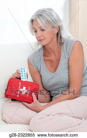 Stock Photography - Beautiful mature woman sitting on her bed and taking tablets out of a red first aid kit. Fotosearch