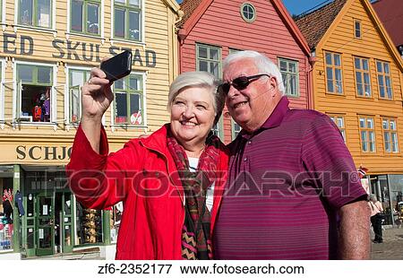 Bergen Norway Bryggen retired tourist couple taking photo in front of colorful old wooden houses and smiling selfie MR-11. View Large Photo Image Stock Photo - Bergen Norway Bryggen retired tourist couple taking photo in front of colorful old wooden houses and smiling selfie MR-11.. Fotosearch