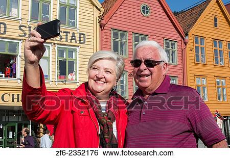Stock Photograph - Bergen Norway Bryggen retired tourist couple taking photo in front of colorful old wooden houses and smiling selfie MR-11.. Fotosearch