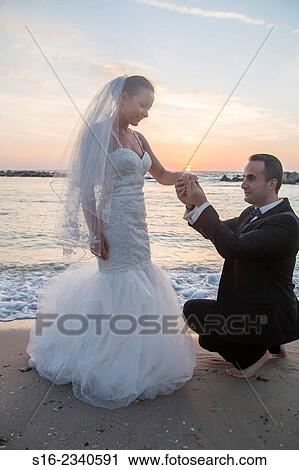 Stock Image - Bridegroom puting a ring on his bride's finger at sunrise on the beach.. Fotosearch