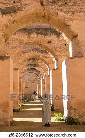Stock Image - Meknes Morocco columns of Hri Souani former horse stalls in 17th century with arab man in robe in downtown with sunlight.. Fotosearch
