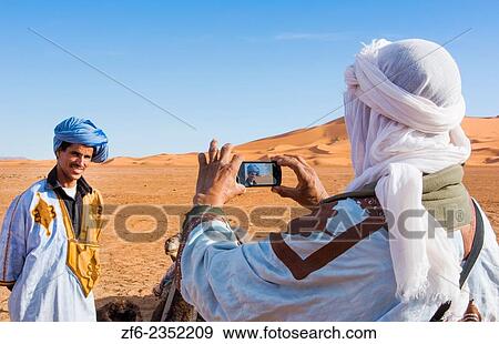 Morocco Sahara Desert sand dunes in Las Palmeras area with local man taking cell phone phoyo of his friend and peaks and sand. View Large Photo Image Stock Photo - Morocco Sahara Desert sand dunes in Las Palmeras area with local man taking cell phone phoyo of his friend and peaks and sand.. Fotosearch