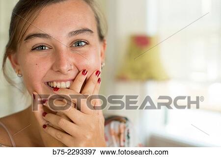 Stock Photo - Portrait of young girl with blue eyes and a nose piercing, looking at the camera with her hands on her chin, smiling and joyful expression of surprise. Fotosearch