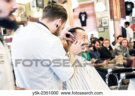 Stock Image - Rotterdam, Netherlands. Barber store Schorem. Barber helping his customer in his barber shop by cutting and modeling his hair.. Fotosearch