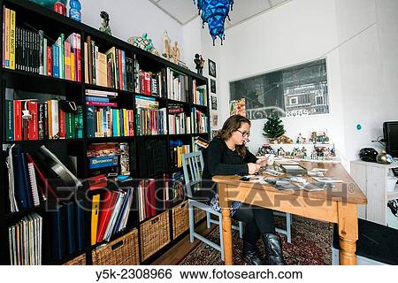 Stock Photograph - Tilburg, Netherlands. Young, single, and thoughtful woman with her library of books and collection of old black and white photographs in her living room.. Fotosearch
