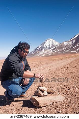 Stock Photo - "Chilean man wearing glacier glasses stacks rocks to create an apacheta in front of Licancabur Volcano, San Pedro de Atacama Desert, Chile, South America. ". Fotosearch