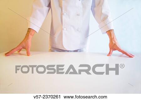 Stock Image - "Cook behind a white table, hands on the table. ". Fotosearch